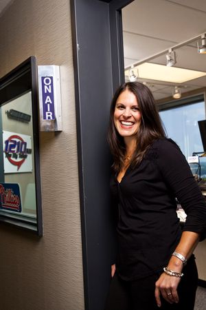 Smiling woman in black outfit standing by an On Air sign at a radio station studio doorway.