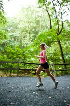 Female runner in pink top and black shorts jogging on a forest trail surrounded by green trees.