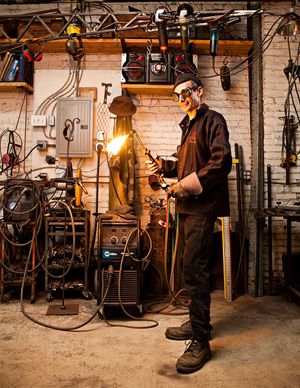 Male welder wearing safety goggles using welding torch in a workshop with tools on brick wall.
