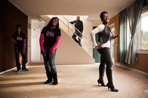 Group of four people posing energetically in an empty carpeted room with stairs in background.