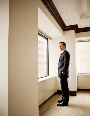 Businessman in dark suit with red tie standing by office window in a corporate hallway.