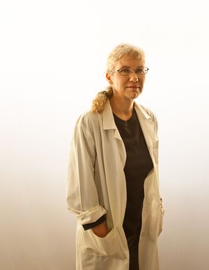 Senior female scientist in white lab coat and glasses standing against a bright white background.