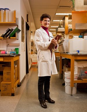 Female scientist in white lab coat smiling while holding a notebook in a research laboratory.