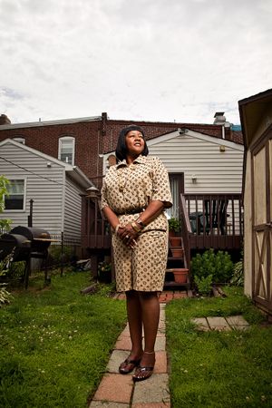 A smiling Black woman in a floral dress stands on a garden path between residential homes.