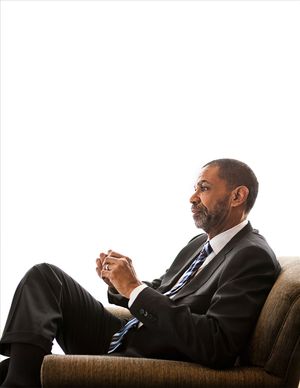 A professional Black man in a dark suit and tie sits thoughtfully in a chair with hands clasped.