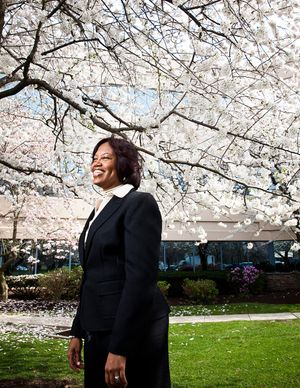 A smiling Black woman in a black suit stands under blooming white cherry blossom trees.