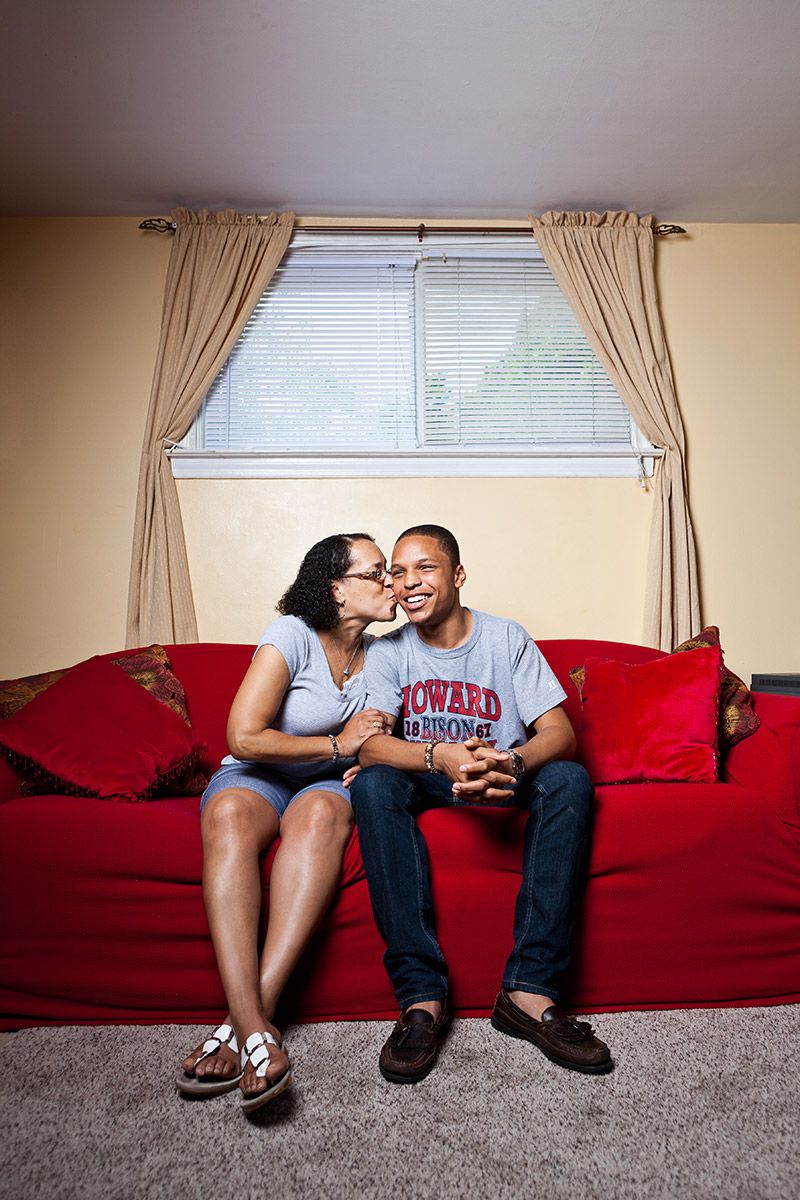 A woman kisses a young man wearing a Howard University shirt on a red couch.