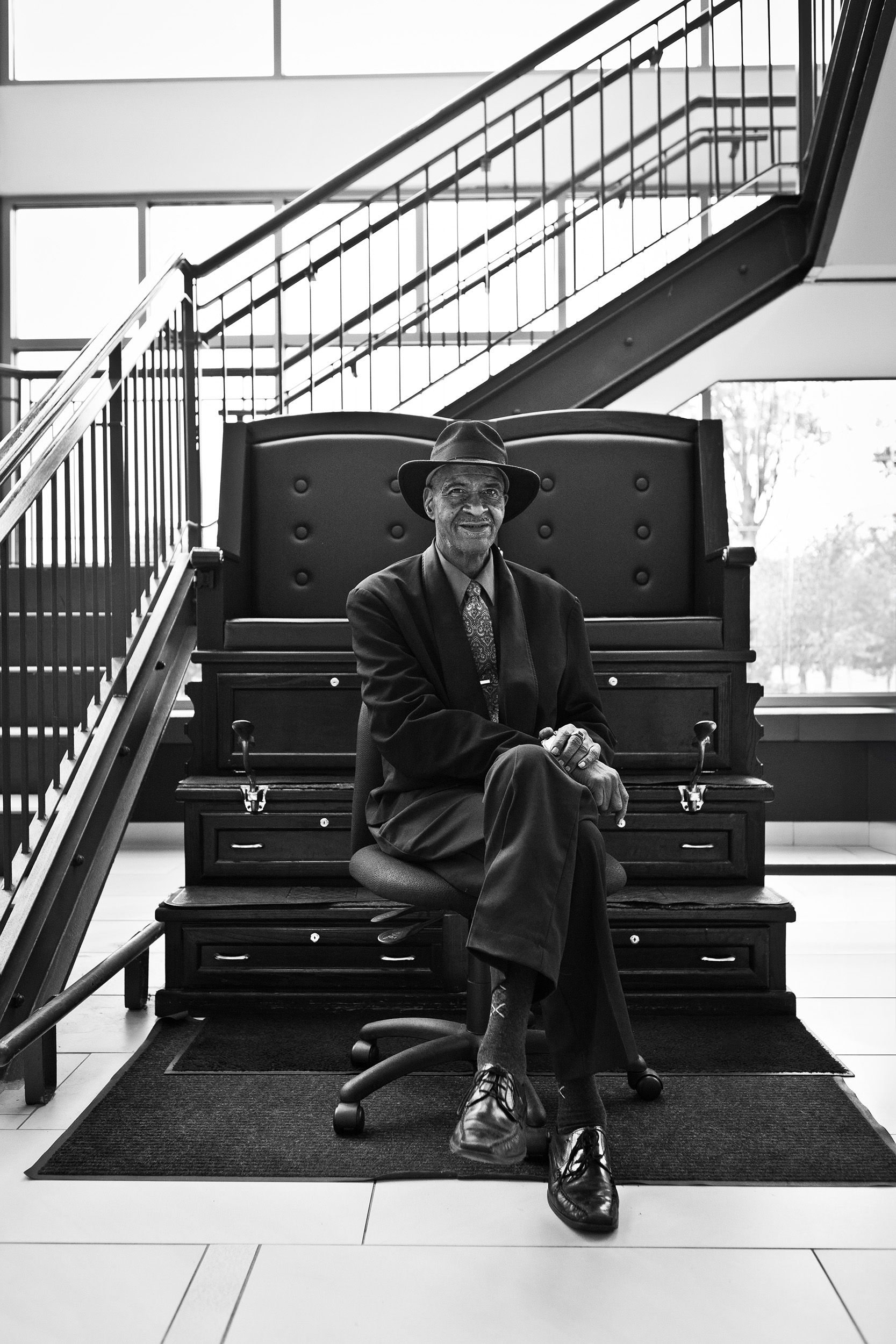 Black and white photo of a man in a hat sitting on a throne-like chair by stairs.