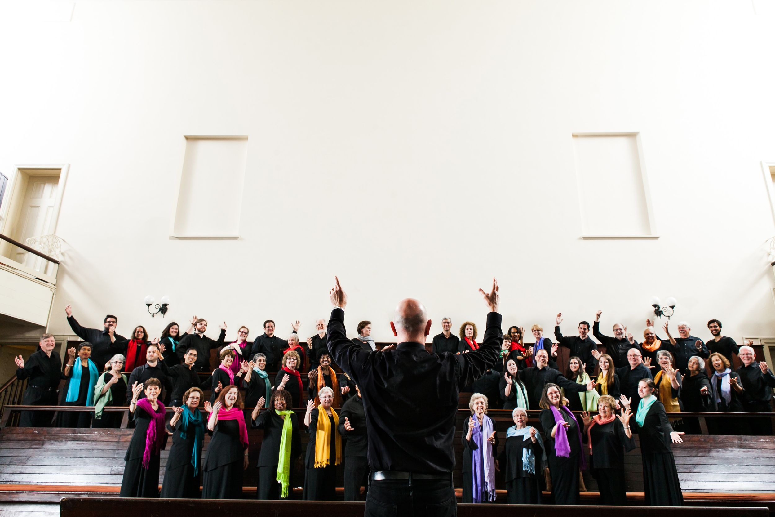Choir conductor leading a large group of singers with raised hands in a bright hall.