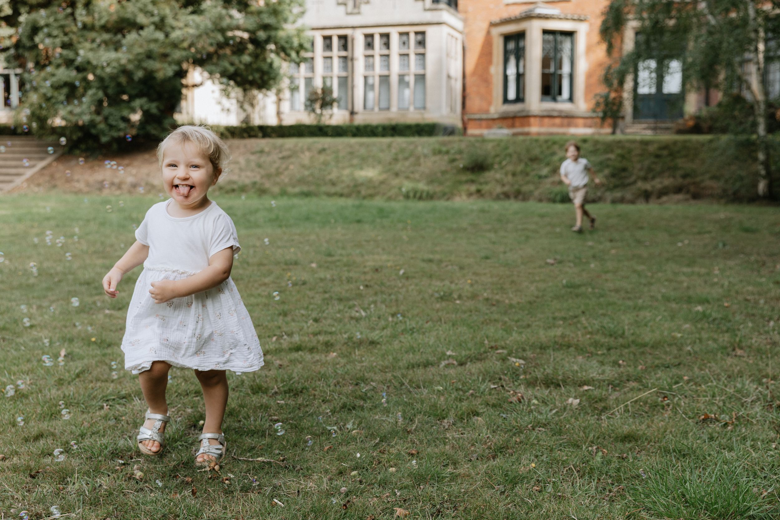 A toddler in a white outfit plays and explores on a grassy lawn near brick buildings.