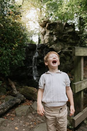 A joyful child wearing a white knit sweater and khaki pants laughs heartily in a garden setting with stone walls.