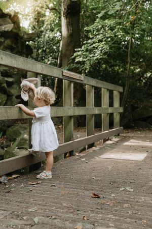 A toddler in a white dress peers through wooden railings on a rustic bridge walkway.