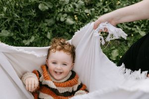 A child in a striped orange sweater lies on a white blanket outdoors with greenery in the background.