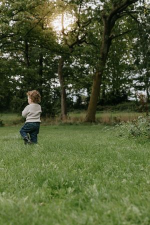 Small child in a sweater and jeans stands in a grassy field surrounded by tall trees at sunset.
