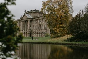 Historic Lyme Park mansion reflected in a tranquil lake on an autumn day with golden trees framing the view.