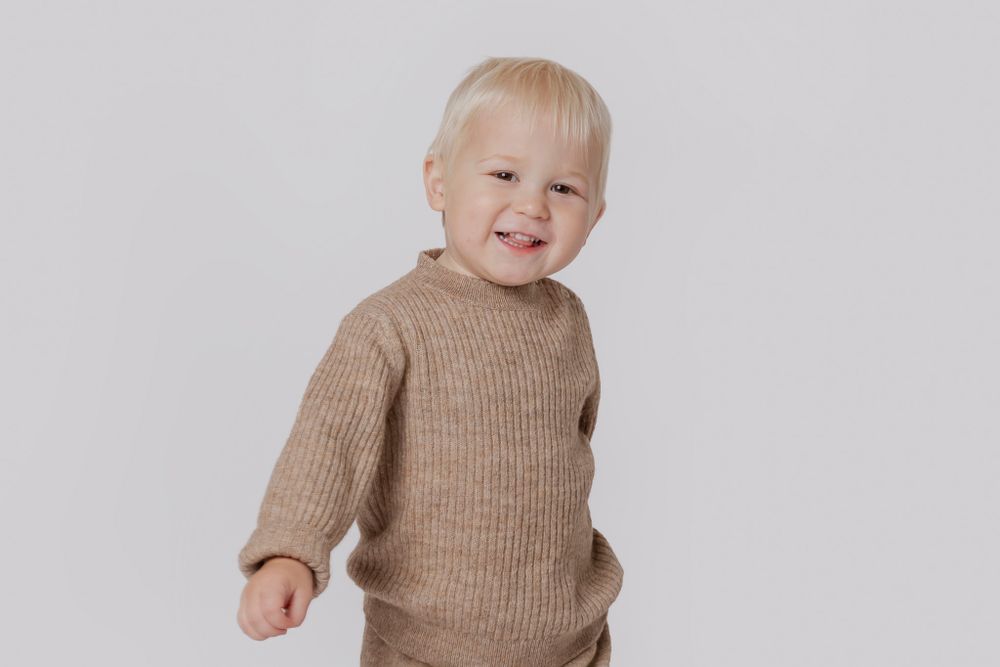 Baby iført brun strikket trøje ligger på hvidt tæppe med glædesfuldt udtryk. Børnefotograf Fyn