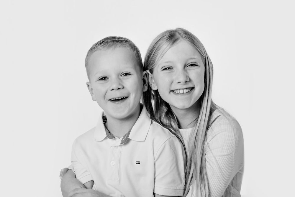 Sort-hvidt studiefoto af to børn, der omfavner hinanden og smiler til kameraet. Børnefotograf Fyn