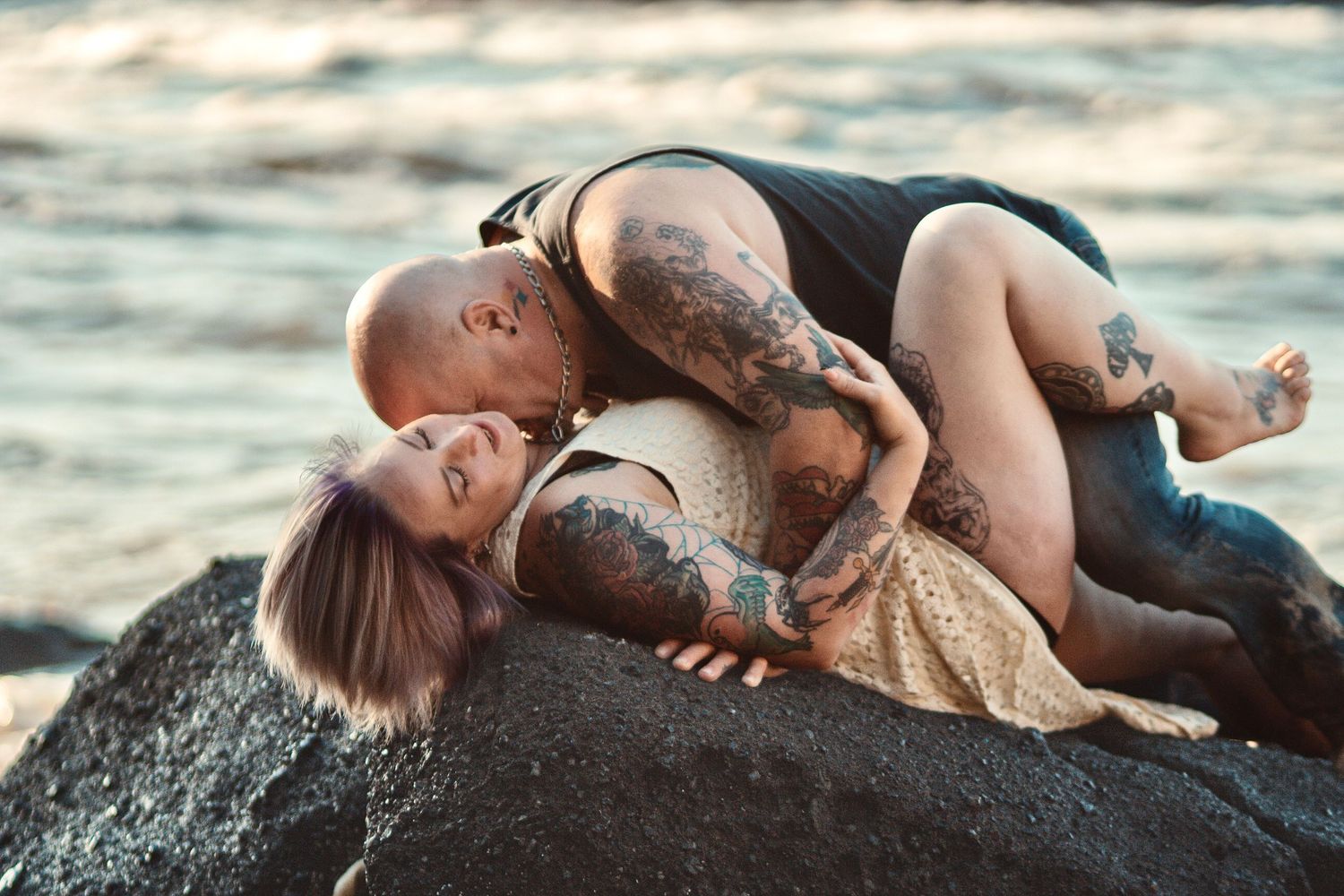 Tattooed couple sharing an intimate moment on coastal rocks with ocean waves in the background.