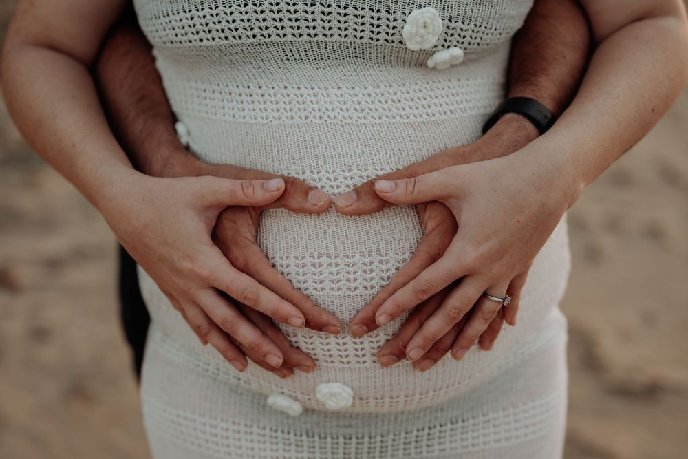 Close-up of hands cradling pregnant belly in white knit dress against gladstone beach backdrop.