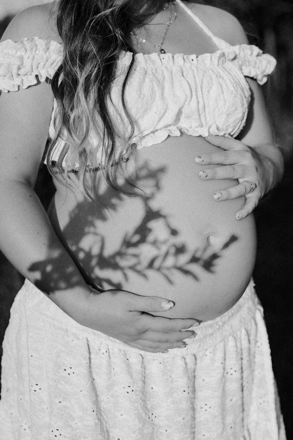 Black and white maternity photo of pregnant woman in white dress cradling bare baby bump with plant shadow overlay.