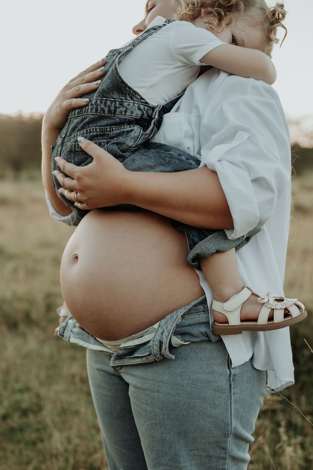 Pregnant woman in white shirt and jeans holding a young child outdoors in a field.