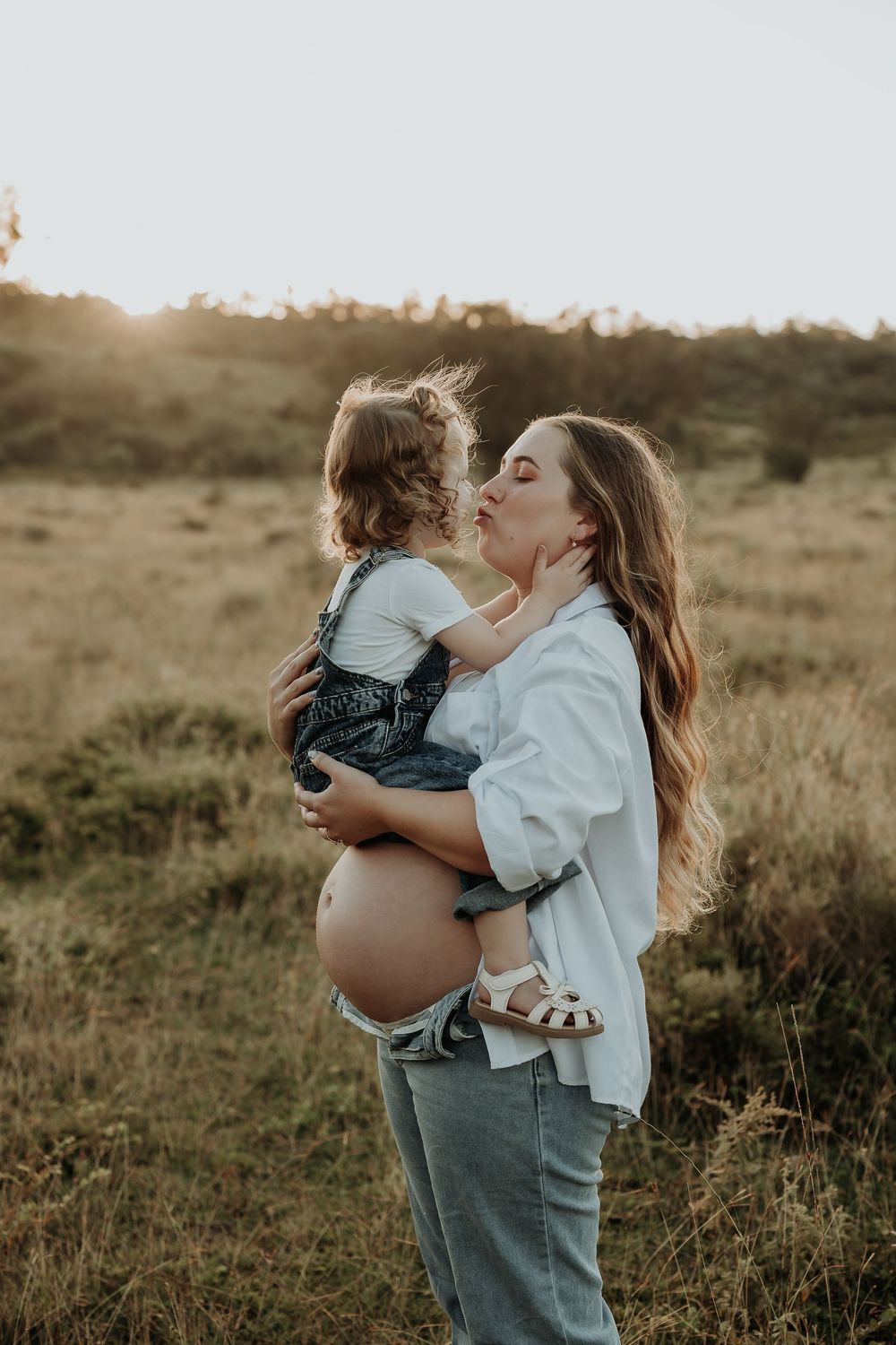 Pregnant woman holding toddler child, sharing a kiss outdoors in golden sunset light in a grassy field.
