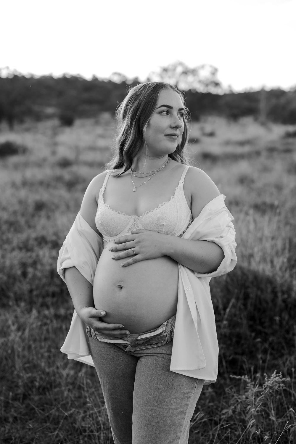 Black and white maternity photo of pregnant woman cradling belly outdoors in field at sunset.