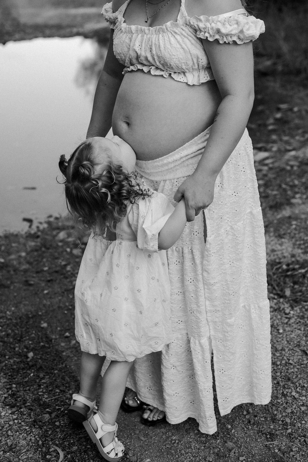 Black and white photo of pregnant woman with toddler girl in white dress hugging her baby bump outdoors.