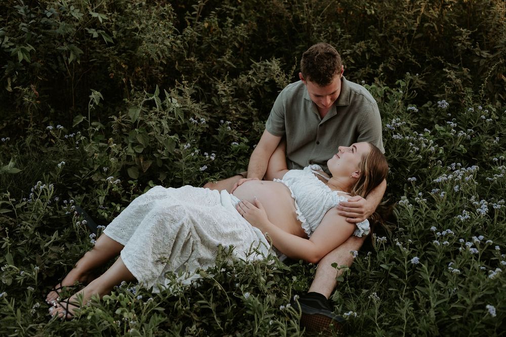 Couple relaxing in a lush green field, woman lying down smiling up at man during outdoor maternity photo session.