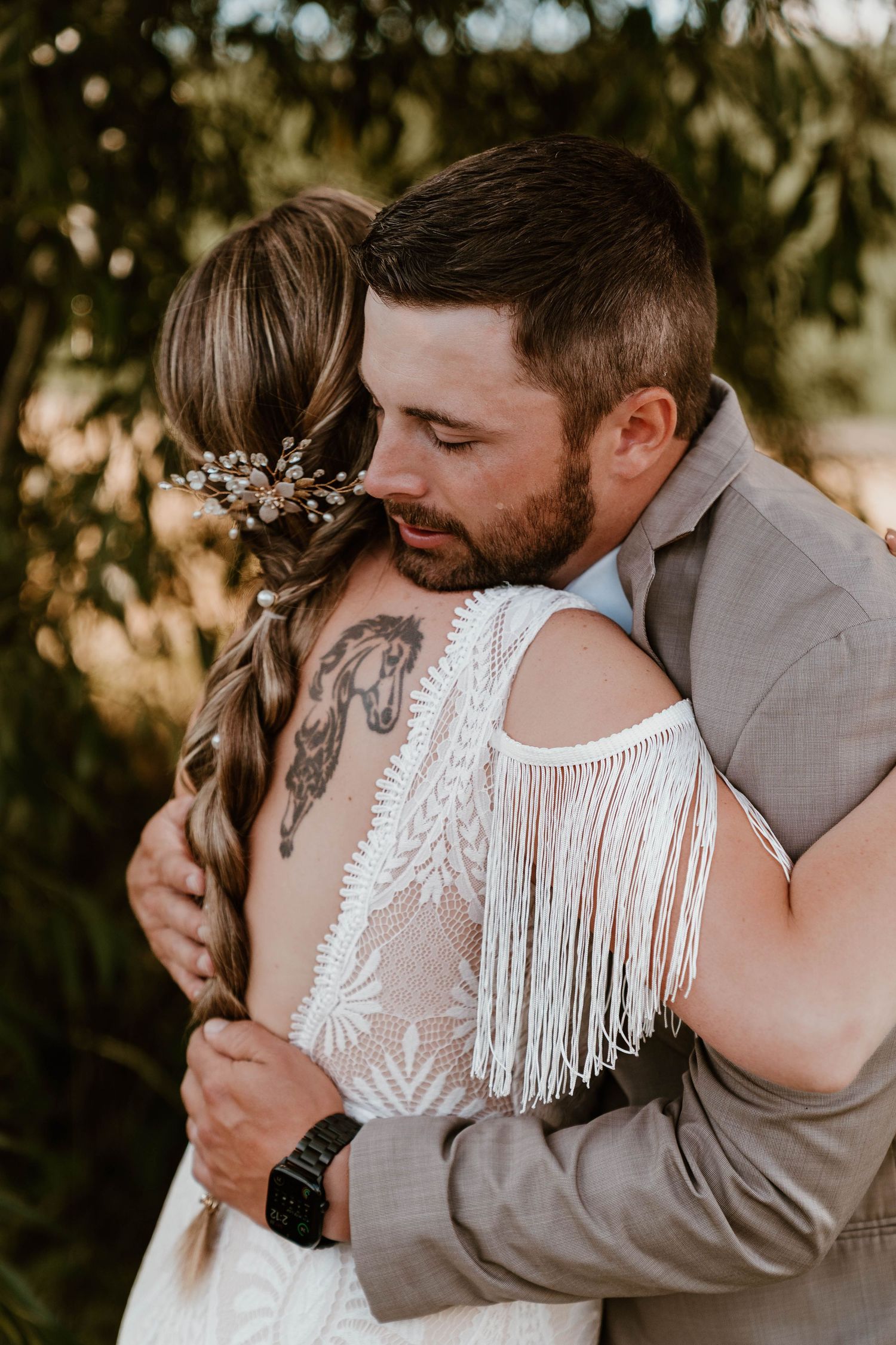 Emotional groom in gray suit embracing bride in boho fringe wedding dress during outdoor ceremony.