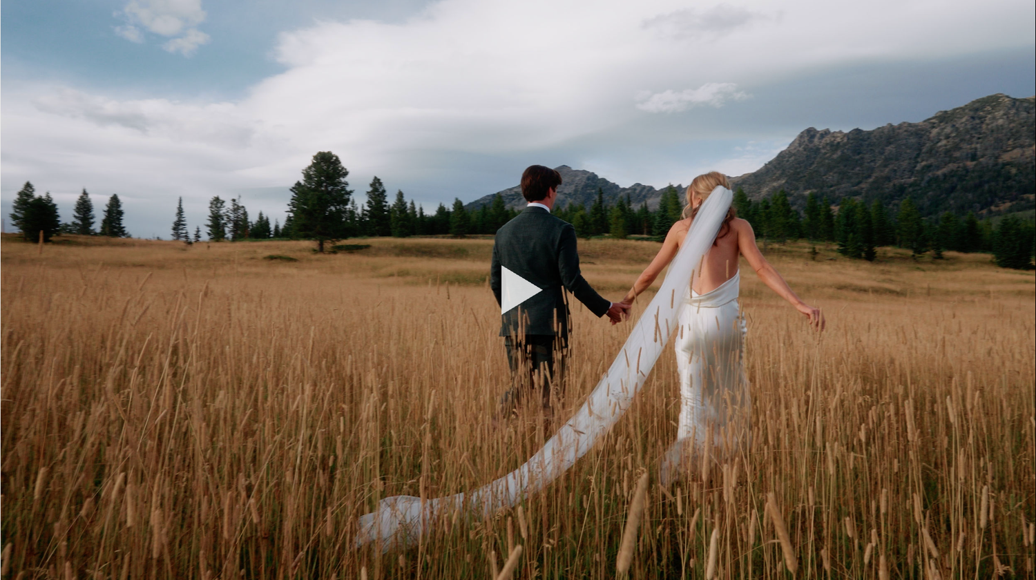 A panoramic view of a rustic barn wedding ceremony setup in a golden wheat field with mountain backdrop.