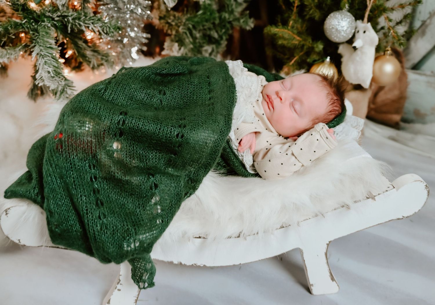 A peaceful sleeping infant wrapped in a green blanket on a white surface with Christmas decor nearby.