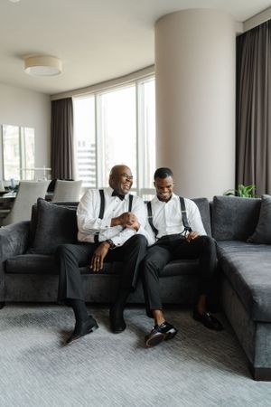 Two men in formal attire elegantly lounging on a modern gray sectional sofa in a bright living room.