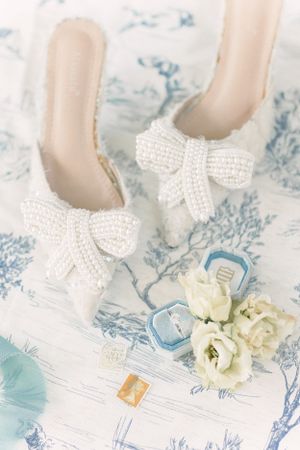 White beaded bow wedding shoes with roses and rings on blue toile fabric.