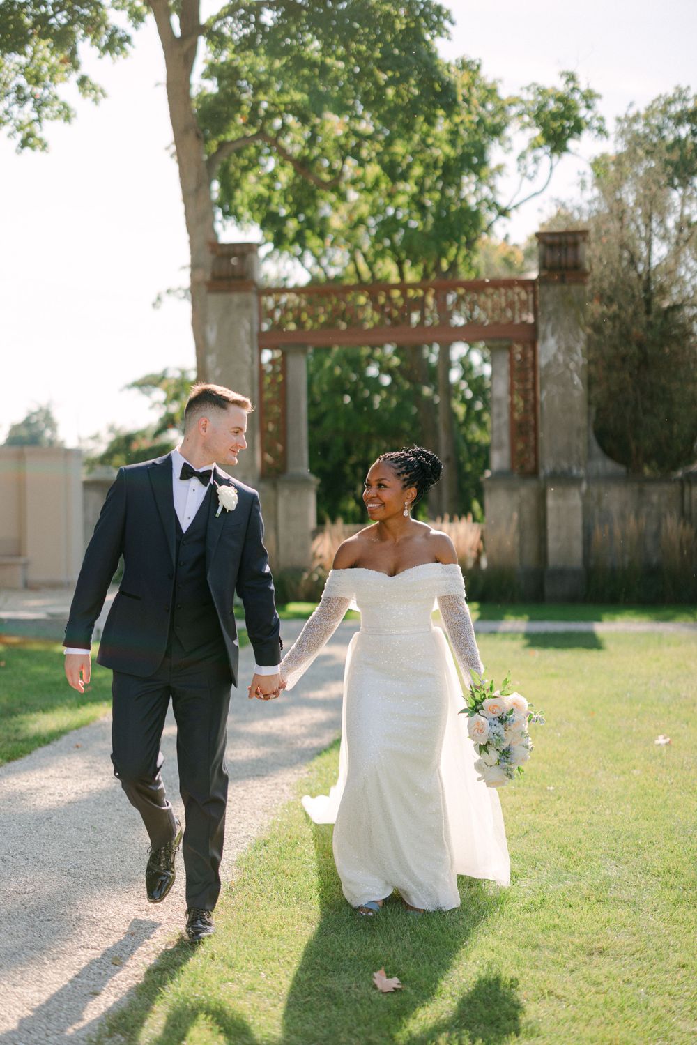 Newlyweds walking hand in hand along a garden path with a rustic arch in the background.