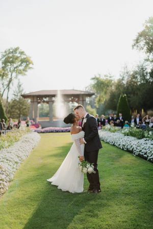 A romantic wedding couple embracing on manicured lawn with fountain and garden backdrop.