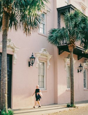 Person walking past pink Charleston building with palm trees and classic architecture.