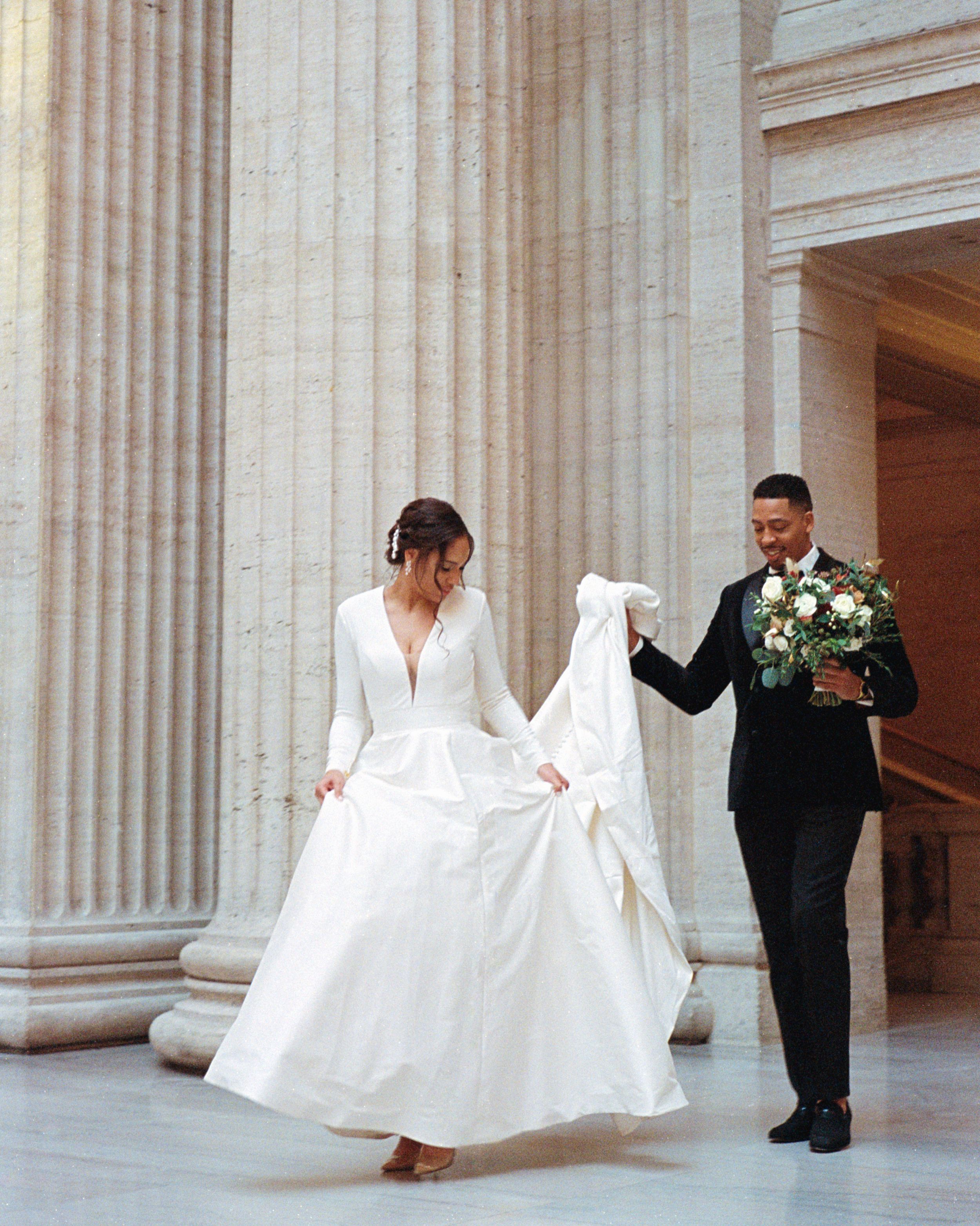 A couple poses elegantly in formal wedding attire between classical stone columns.
