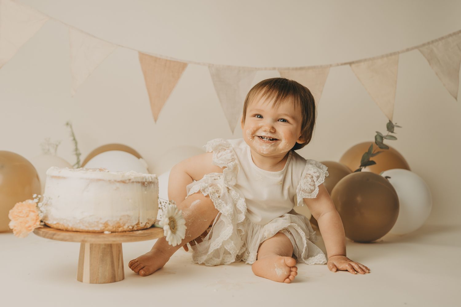 Baby in pink outfit sits among rustic cake smash setup with floral accents.