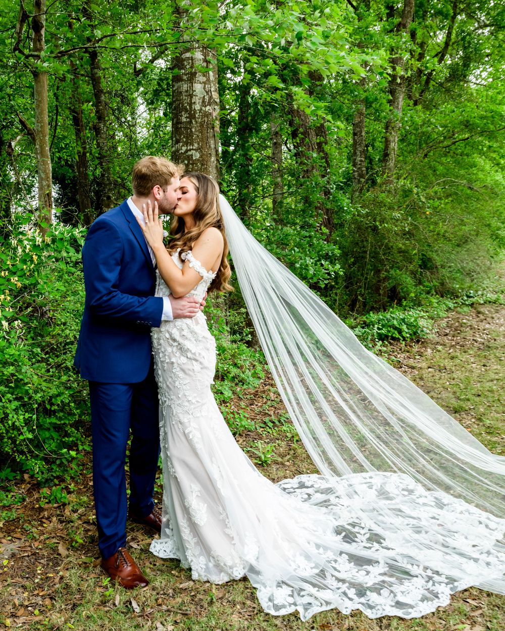 A couple shares an intimate moment in a lush forest setting, with the bride wearing a flowing lace gown and long cathedral veil.