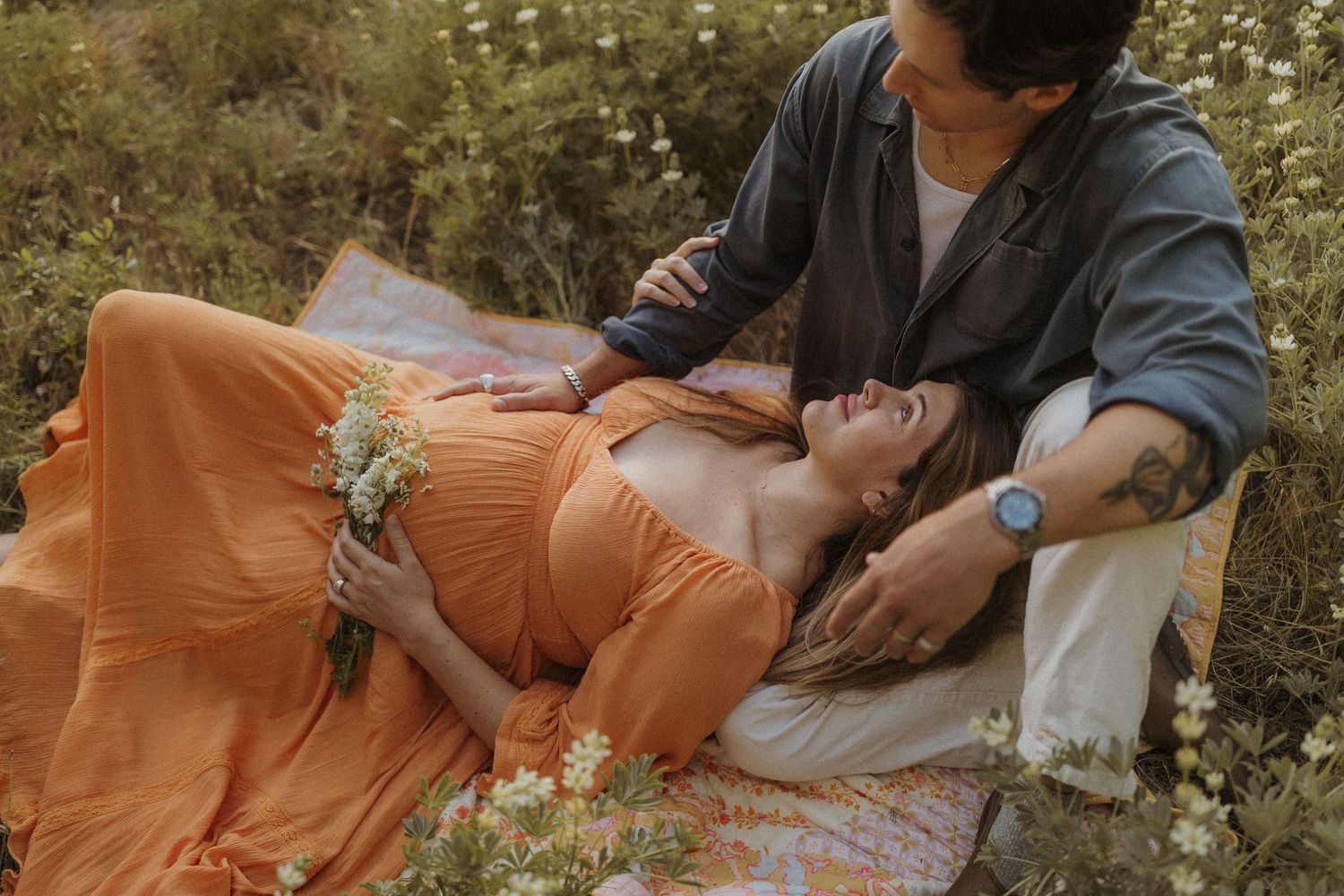 A couple relaxes on a picnic blanket surrounded by white wildflowers in a romantic outdoor setting. San Diego Maternity Photography.