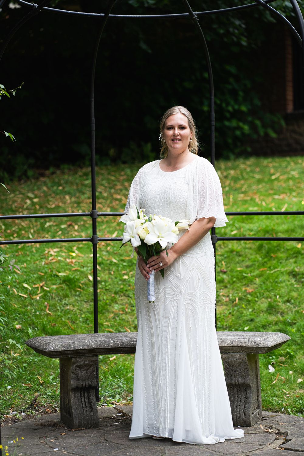 A bride in a flowing white dress poses by a rustic stone bench in a lush garden setting.
