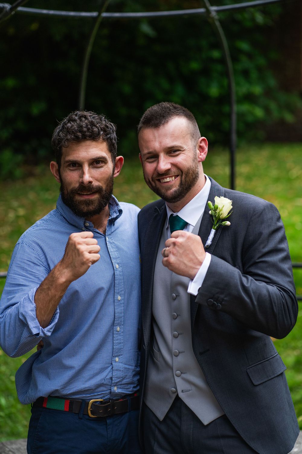 Two men in formal attire striking playful boxing poses together against a green outdoor background.