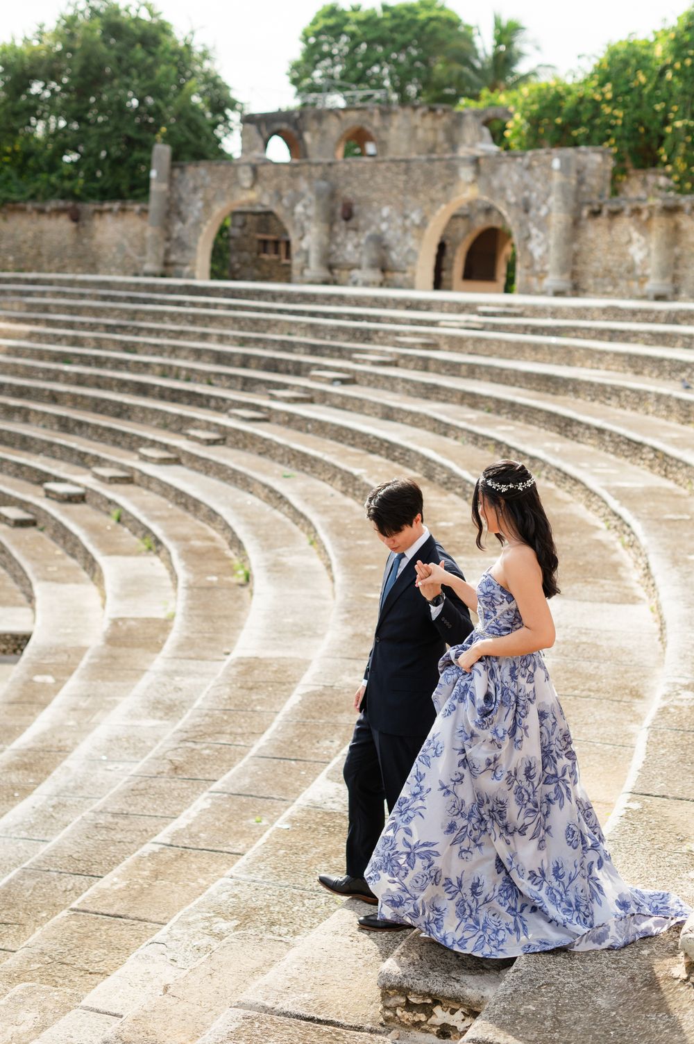A couple sits together on ancient stone amphitheater steps wearing formal attire with a blue and white floral dress.