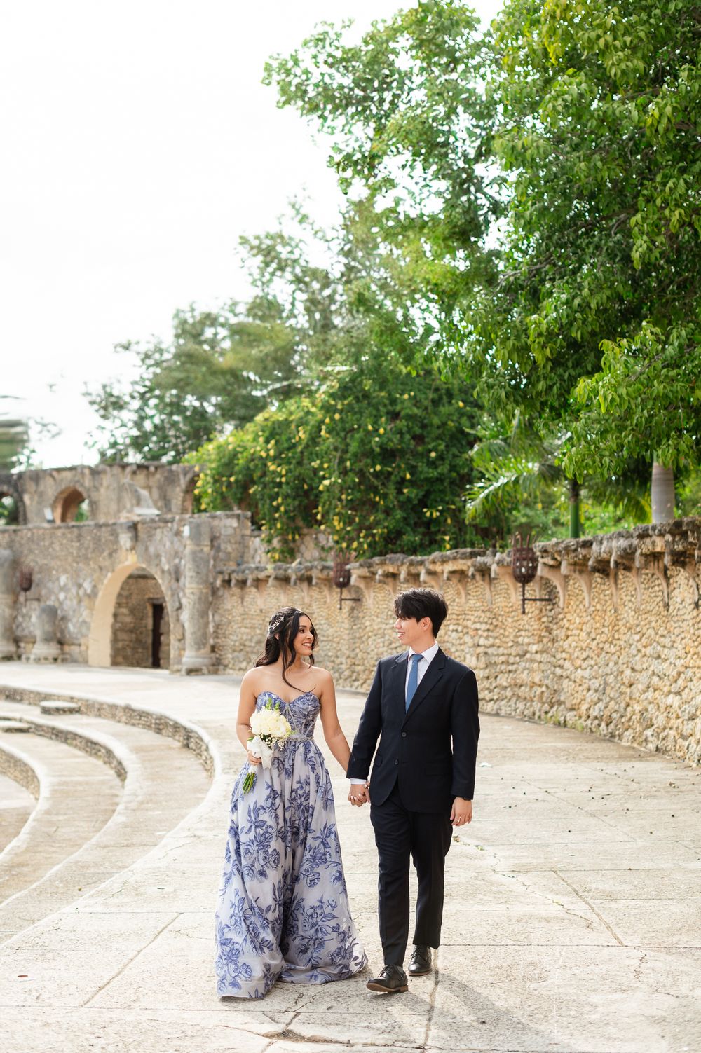 A couple walks together along an ancient stone wall pathway surrounded by lush greenery on a sunny day.