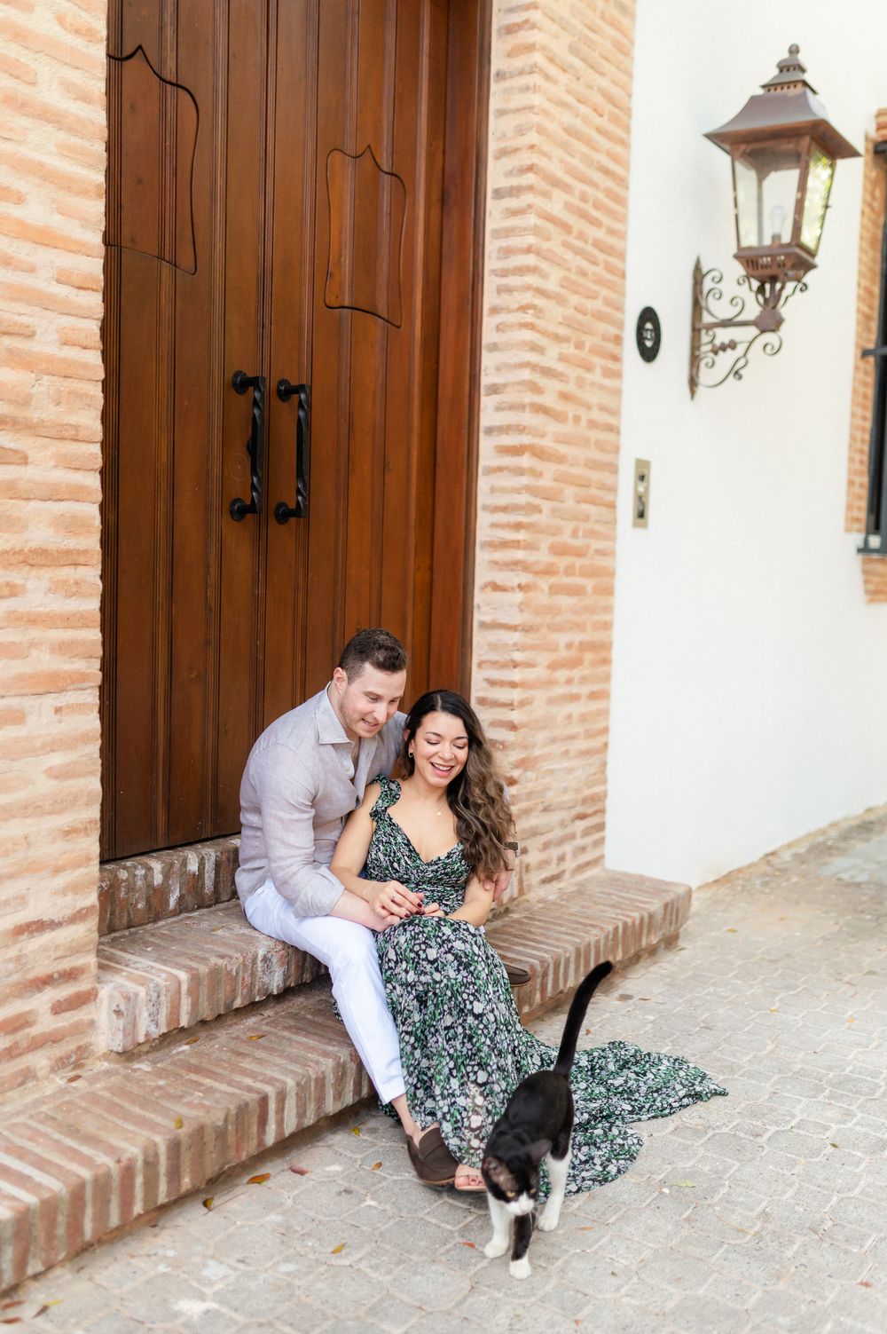 A couple poses playfully in front of rustic wooden doors and brick walls in a romantic outdoor photoshoot.