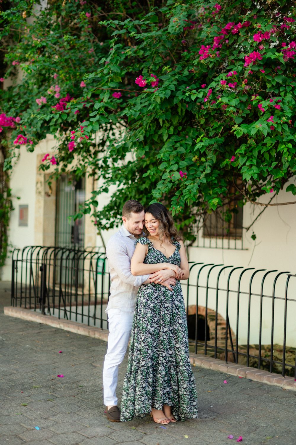 A couple shares a tender moment under flowering bougainvillea along an iron-fenced walkway.