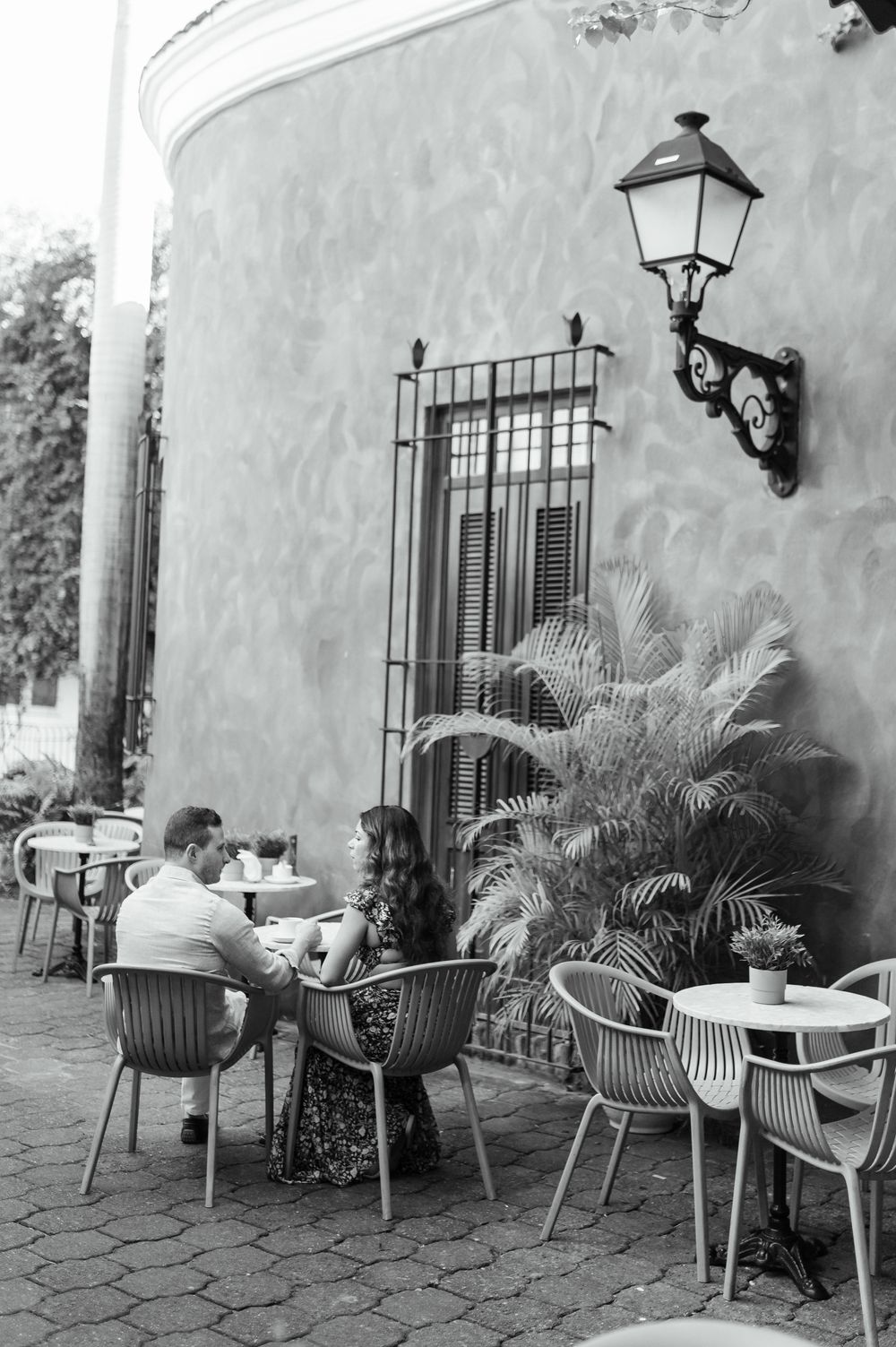 People dining at an outdoor cafe with stone walls, vintage lamp post and palm plants in black and white photography.