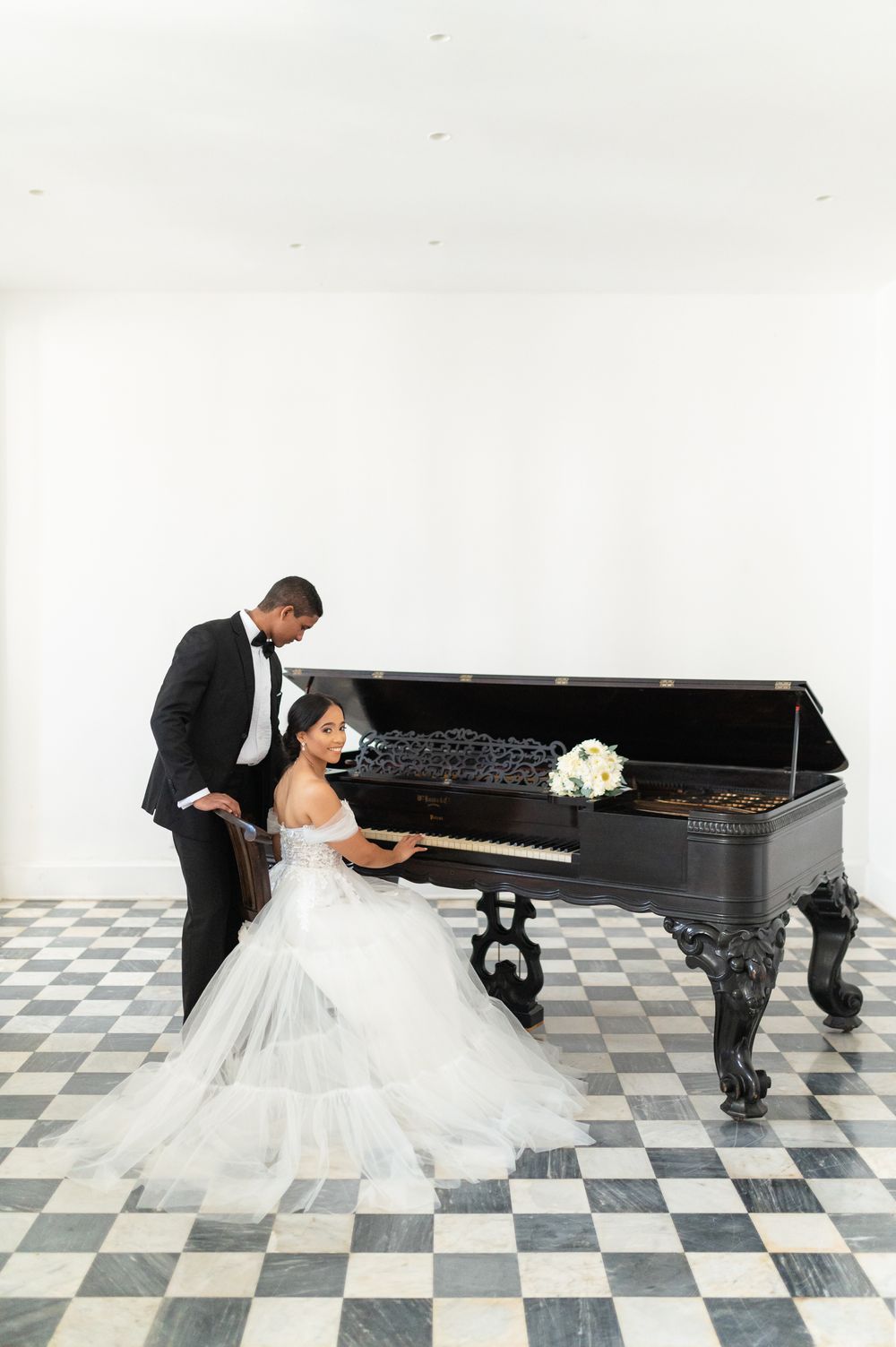 A bride in a white gown and groom in a black suit pose with a grand piano on a checkered floor.