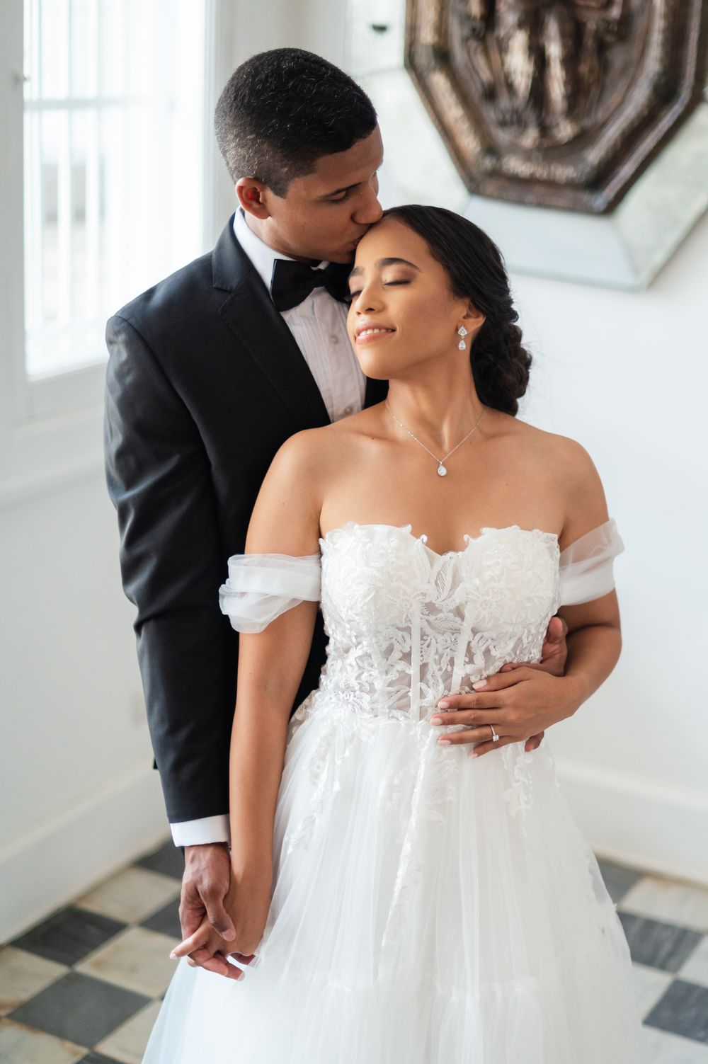 Romantic wedding couple embraces in soft natural light against white walls with elegant decor.